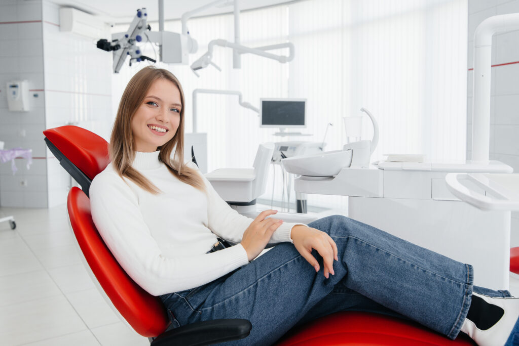 A beautiful young woman is sitting on a red dental chair in modern white dentistry and smiling. Dental prosthetics, treatment and teeth whitening.